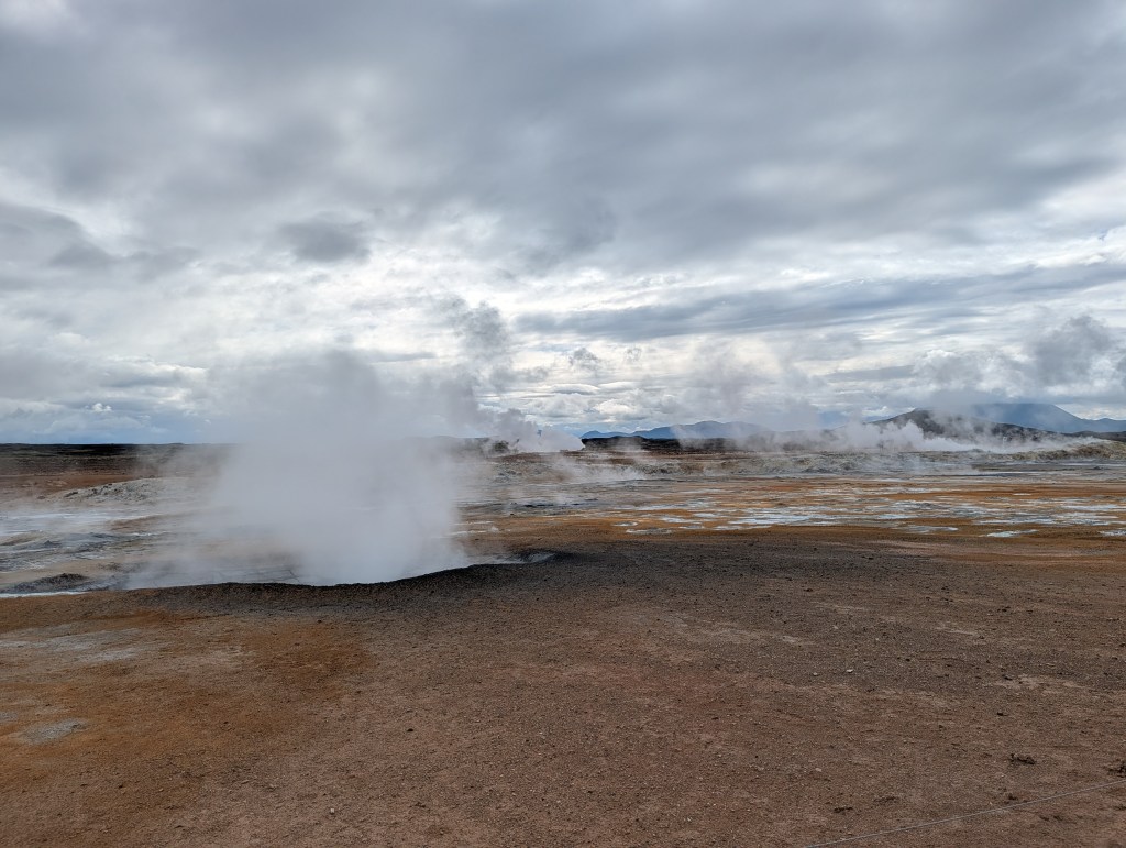 A geothermal landscape with steam rising from multiple vents against a cloudy sky.