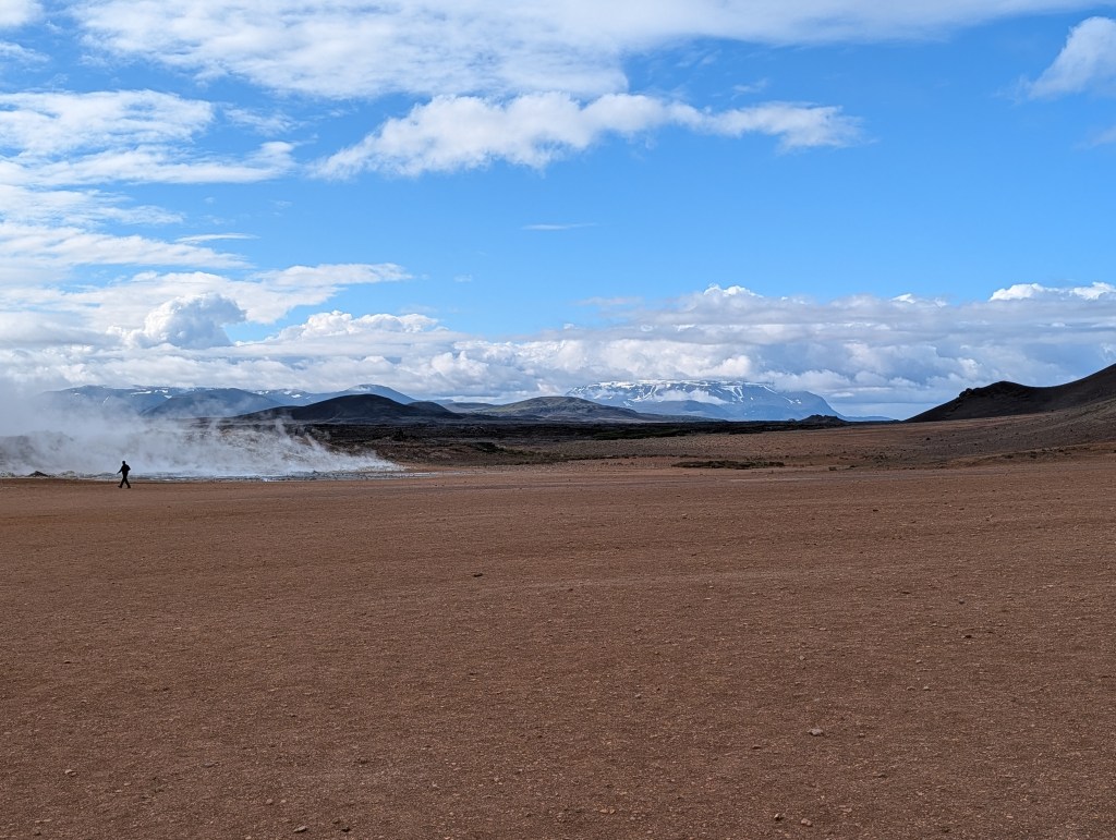 A lone figure walking across a barren landscape with steam rising in the background, under a blue sky with scattered clouds and mountains in the distance.