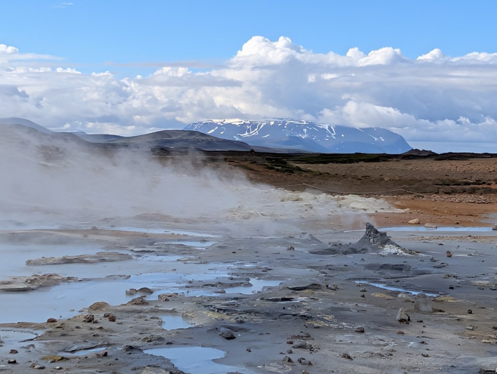 A geothermal area featuring steam rising from hot springs, surrounded by a rocky landscape and distant snowy mountains under a blue sky with scattered clouds.