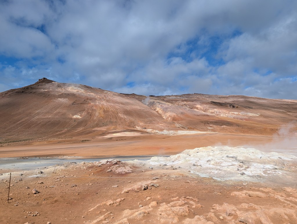 A desolate landscape showcasing arid hills and rocky terrain under a partially cloudy sky, with steam rising from geothermal areas.