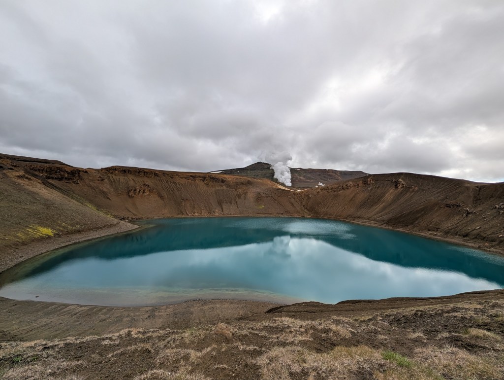 A serene volcanic crater lake with turquoise water surrounded by brown hills and overcast skies. Steam rises from a distant geothermal area in the background.