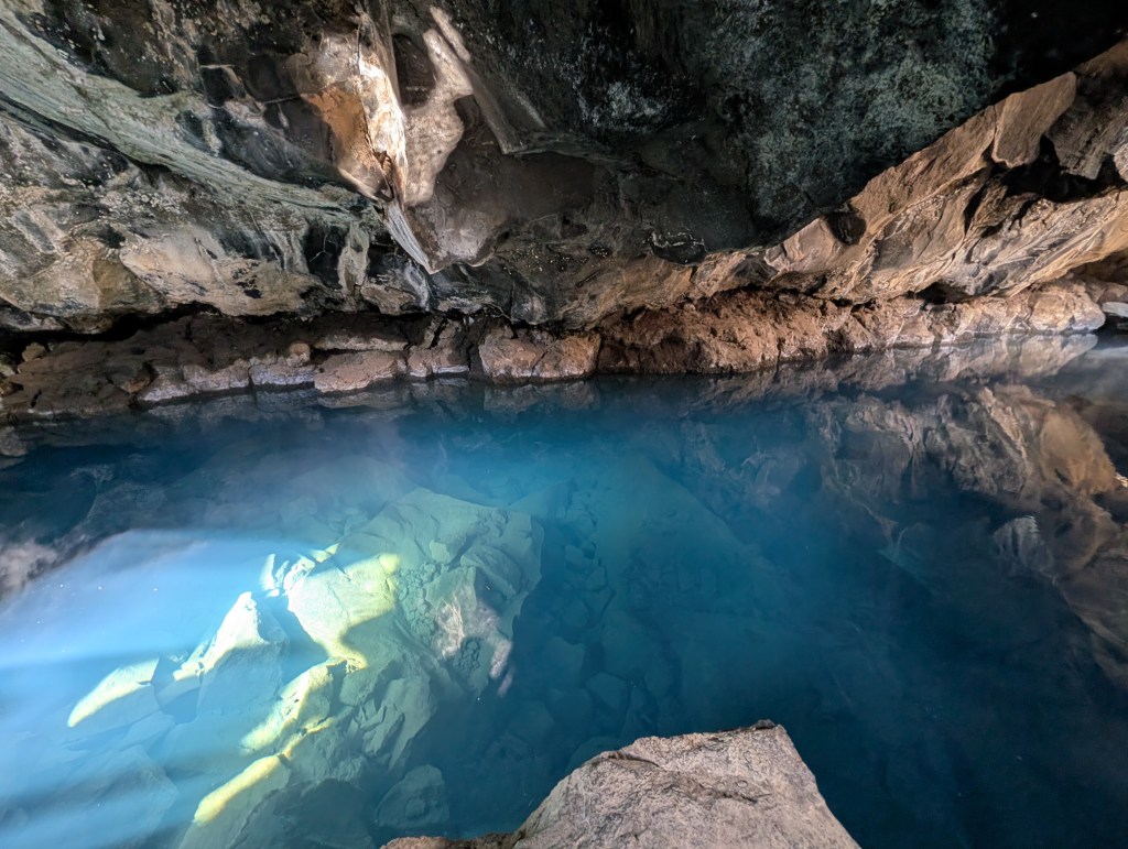 An underground cave featuring a clear blue pool of water, with rocky edges and natural stone formations above.