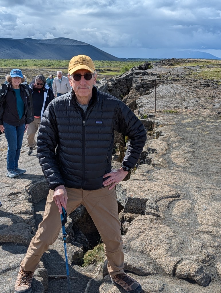 A man in a black puffer jacket and sunglasses stands with his legs apart on rocky terrain, holding a hiking pole. In the background, a group of hikers and mountains under a cloudy sky can be seen.