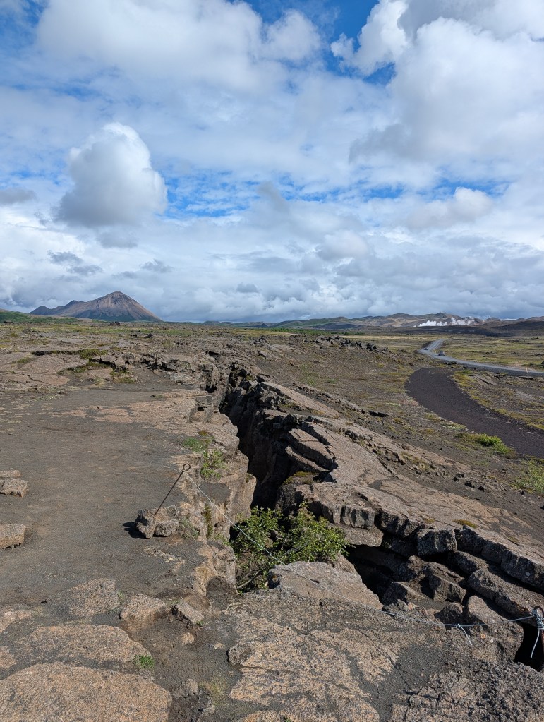 A panoramic view of a rocky landscape with a fissure, surrounded by clouds and distant mountains.