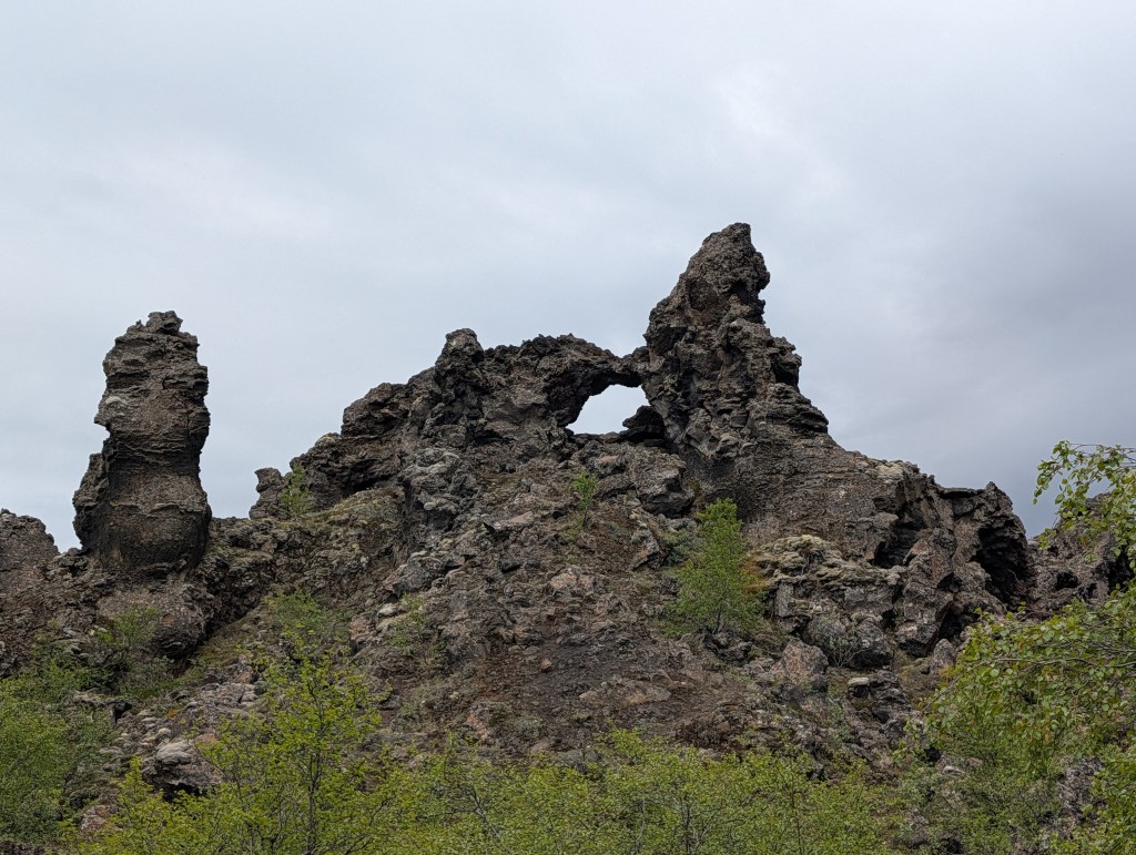 Unique rock formation with a prominent arch, surrounded by greenery and a cloudy sky.
