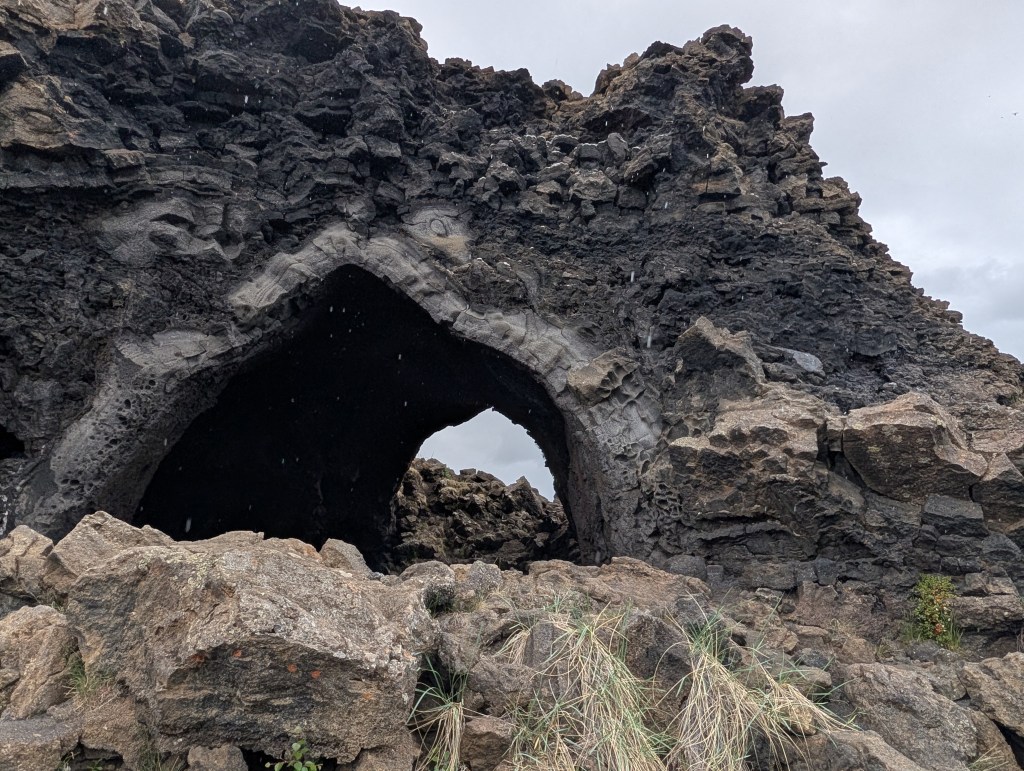 A view of a large volcanic rock formation with a prominent arch, surrounded by smaller rocks and sparse vegetation.