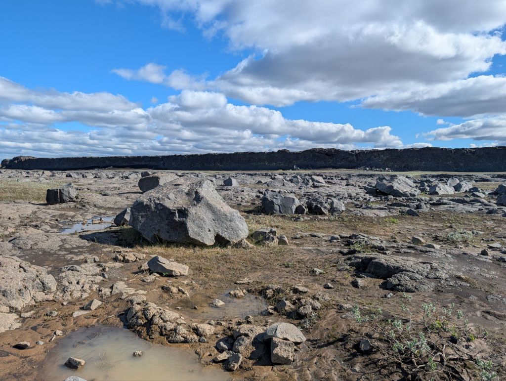 A barren landscape featuring large rocks and patches of grass under a partly cloudy blue sky.