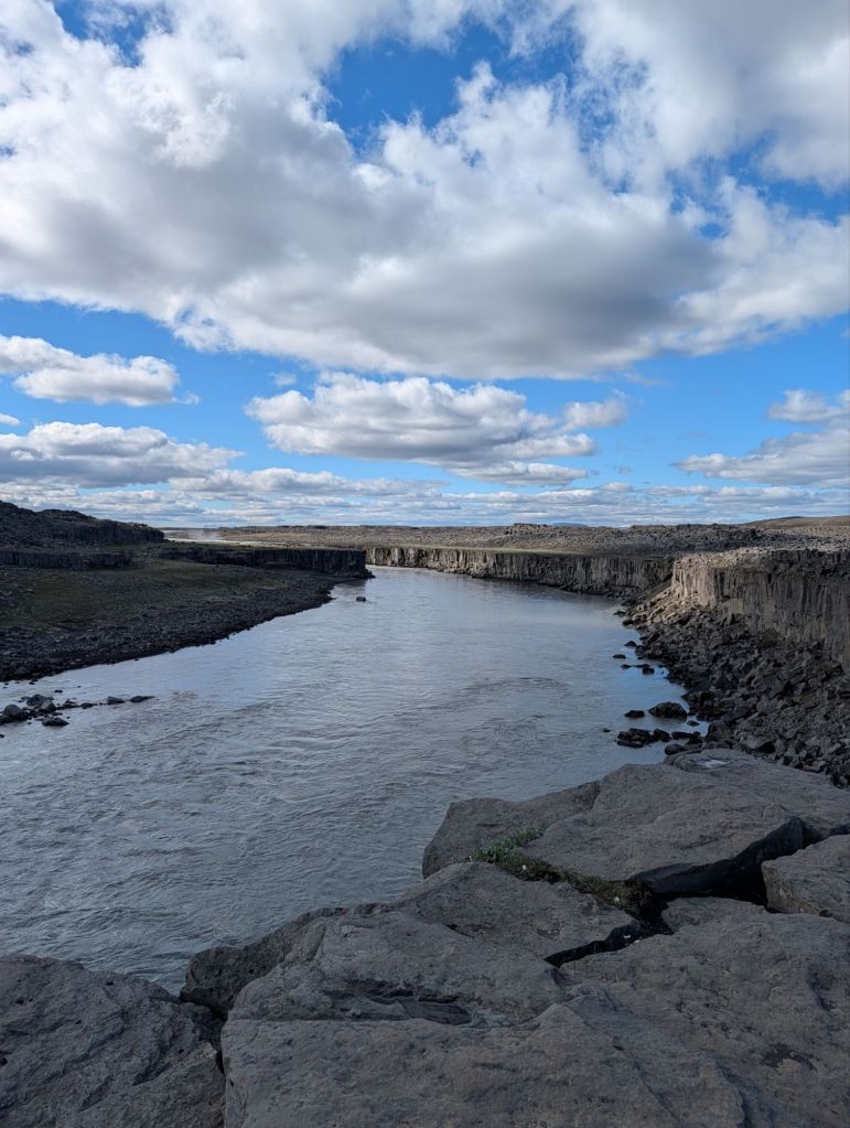 A serene landscape featuring a river flowing through rocky terrain, flanked by tall cliffs under a partly cloudy blue sky.