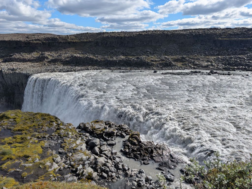 A wide view of a powerful waterfall cascading over rocky cliffs, surrounded by a rugged landscape and dramatic cloud-filled sky.