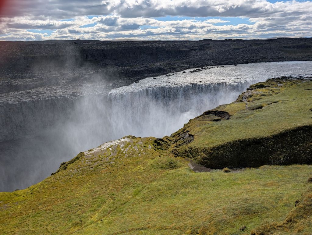 A dramatic view of a waterfall cascading over a cliff, surrounded by rocky terrain and green moss. The sky is partly cloudy with sunlight reflecting off the water mist.
