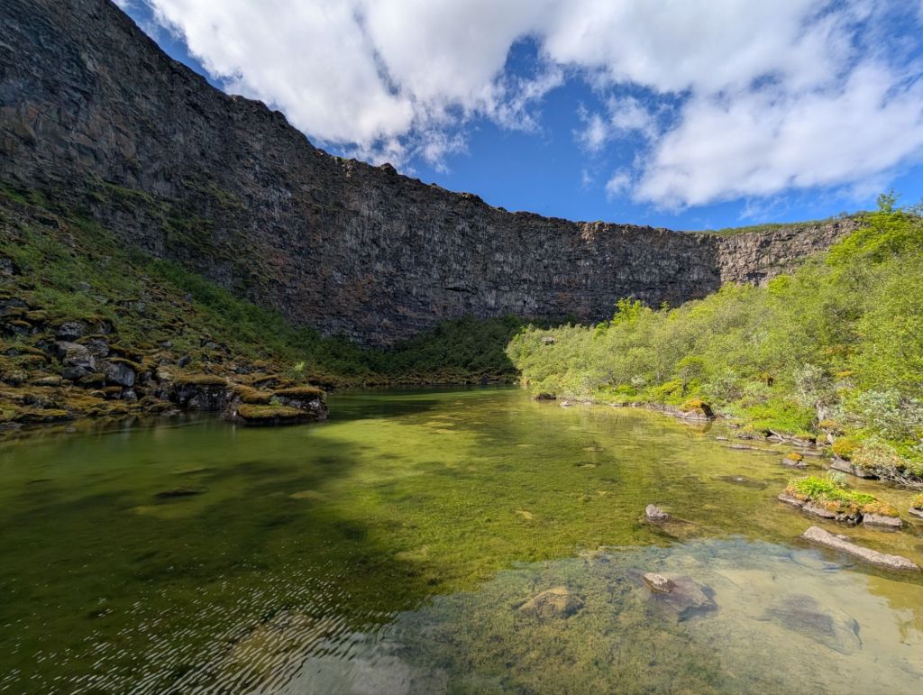 A serene landscape featuring a calm, greenish lake surrounded by rocky cliffs and lush vegetation under a blue sky with clouds.