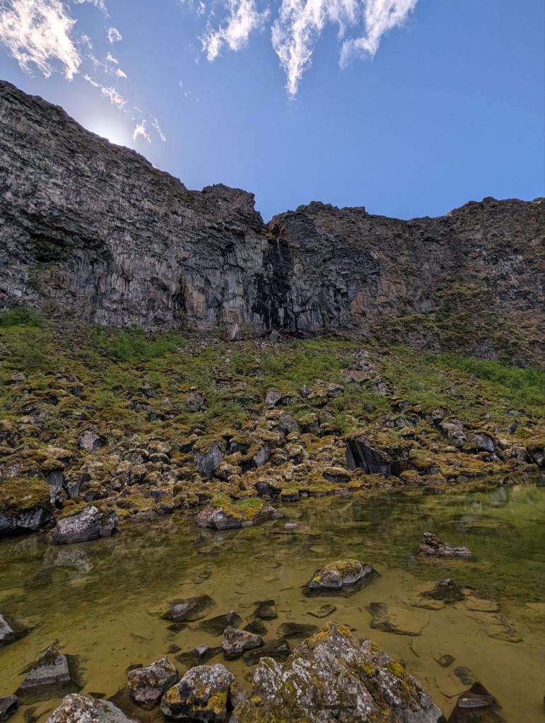 A rocky cliff with a small waterfall, surrounded by green moss and a clear pool of water at the base, under a blue sky with scattered clouds.