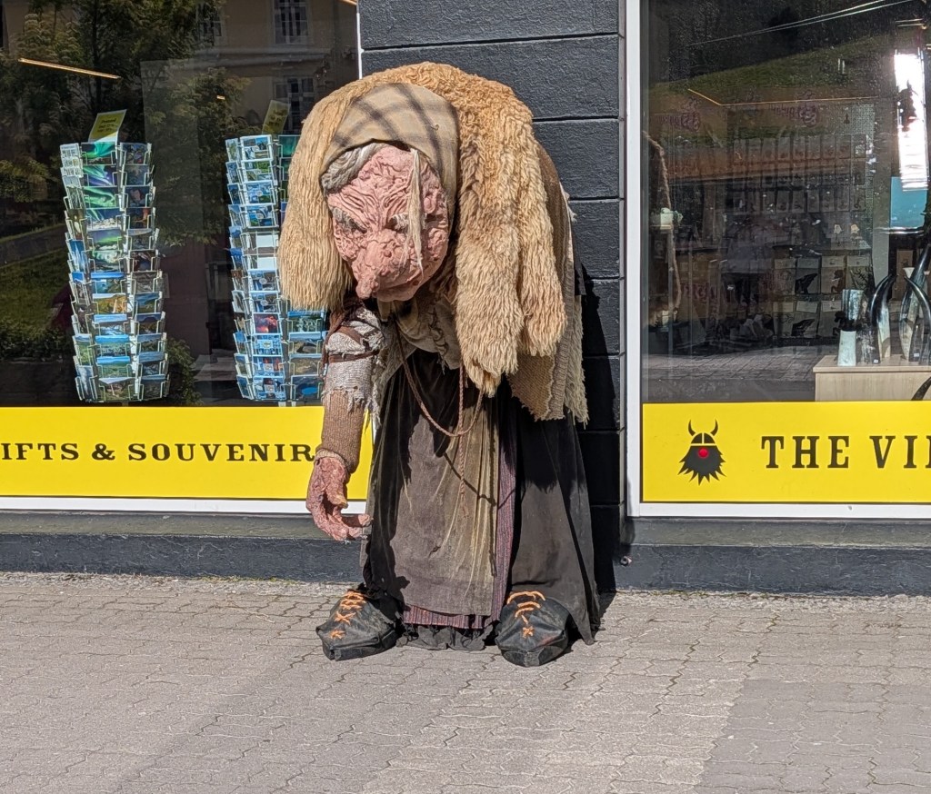 A large, humanoid puppet resembling an elderly woman stands outside a souvenir shop, with a shaggy fur coat and tattered clothing, looking down as if in thought.