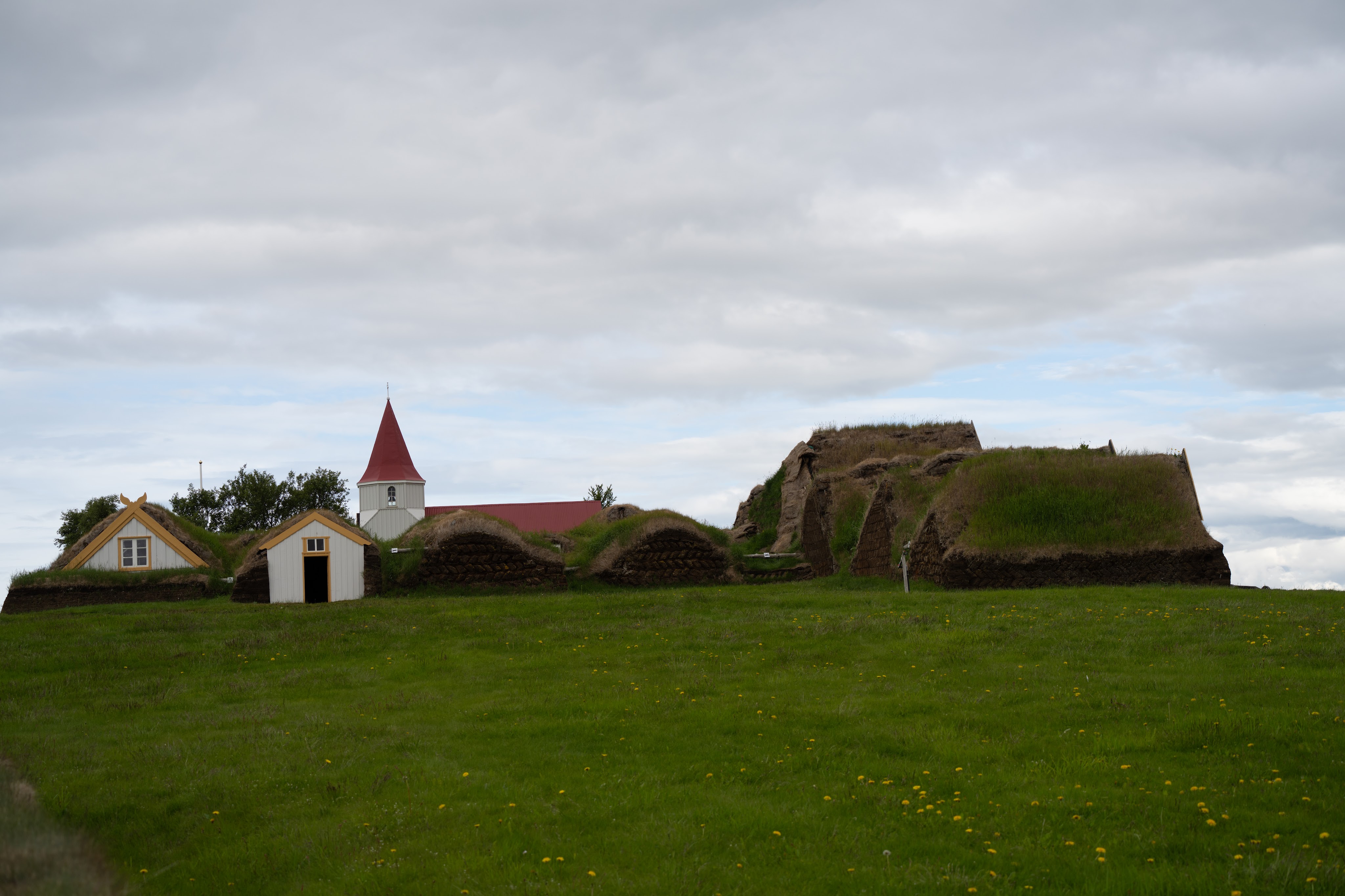 A scenic view of traditional Icelandic turf houses and a church with a red steeple, set against a cloudy sky and green grass.