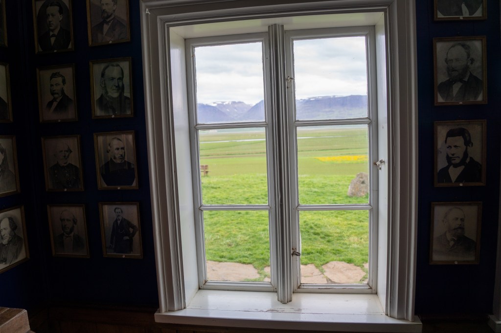 Interior view of a room featuring a window, framed by historical portraits on the walls, overlooking a green landscape with mountains in the distance.