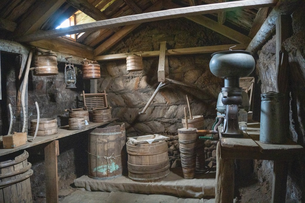 An old rustic interior of a storage room with wooden shelves filled with various kitchen and farm tools, including barrels, a grinder, and lanterns.