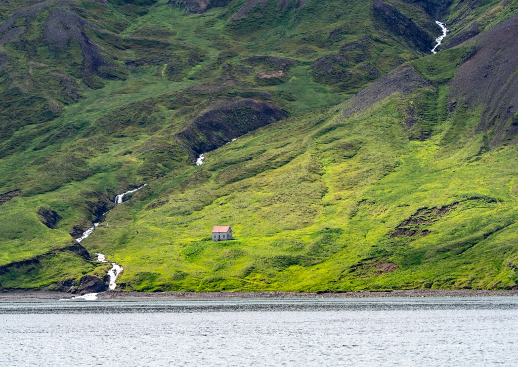 A solitary house surrounded by lush green hills and small waterfalls, viewed from a body of water.
