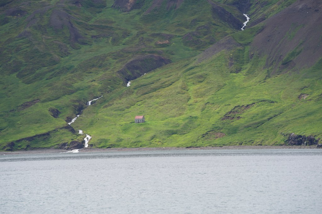 A small house with a red roof situated on a lush green hillside, surrounded by flowing streams, near a body of water.