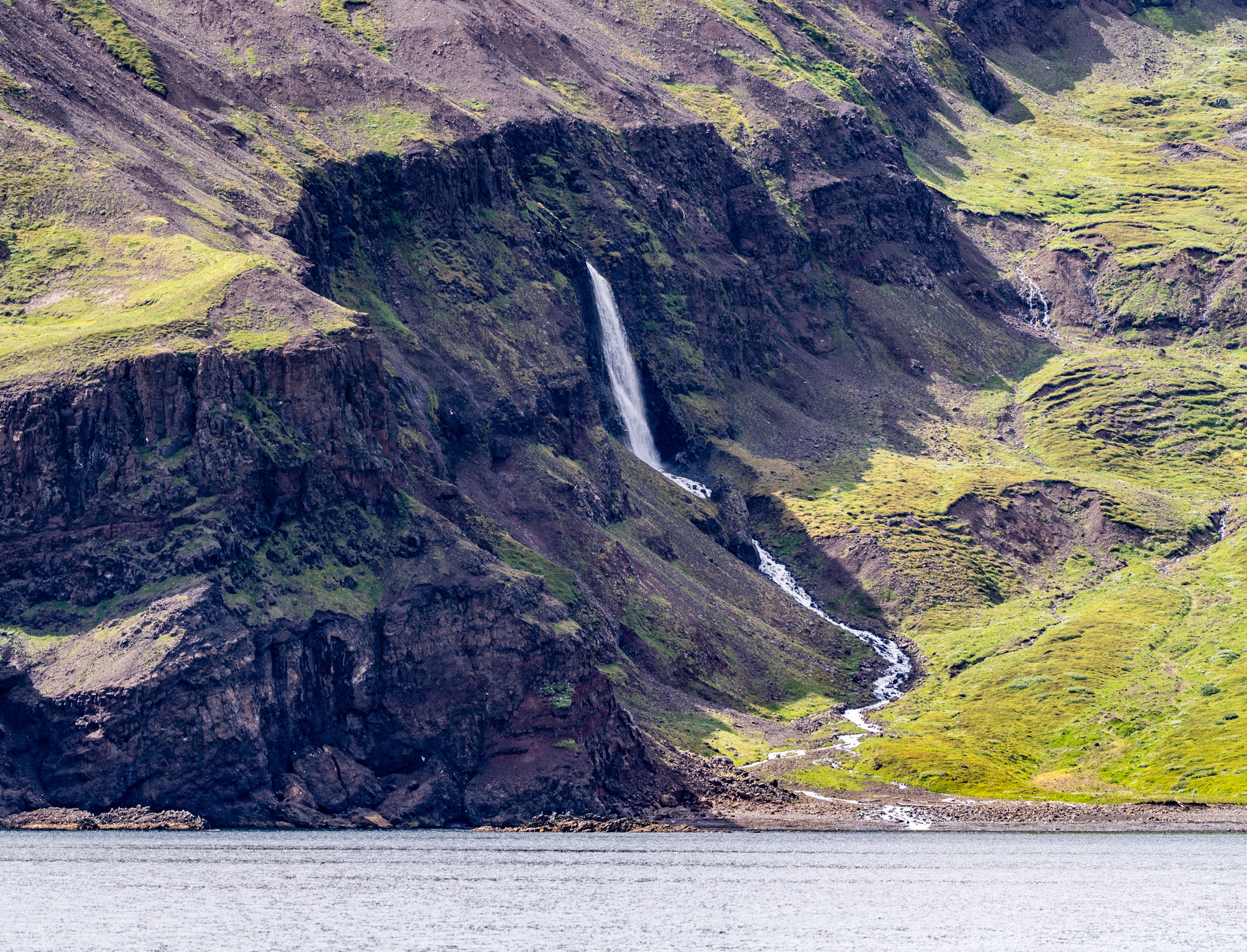 A scenic view of a waterfall cascading down a rocky hillside, surrounded by green grass and rugged terrain, with a calm water body in the foreground.
