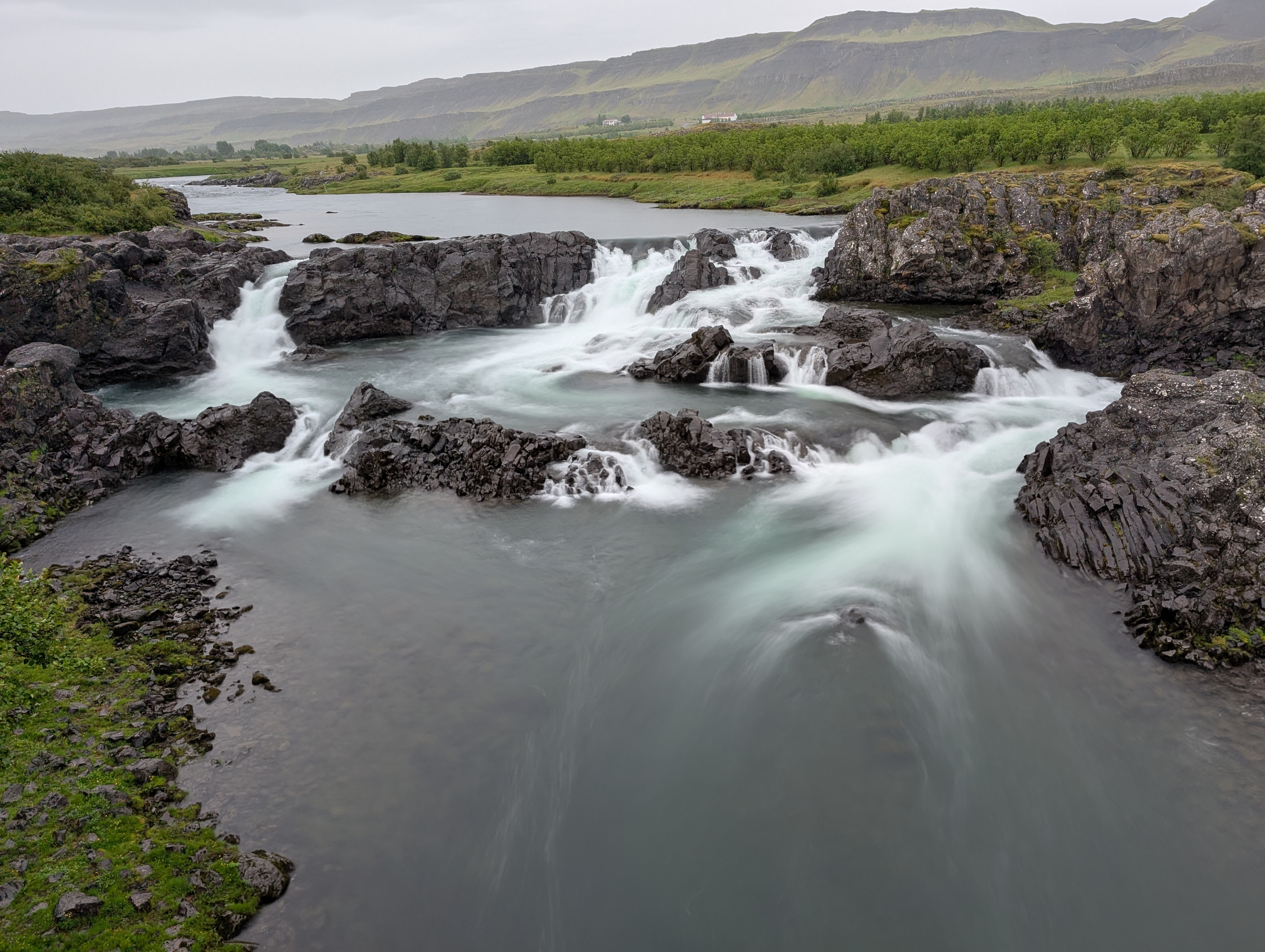 A scenic view of a river cascading over rocky formations, with lush greenery and hills in the background.