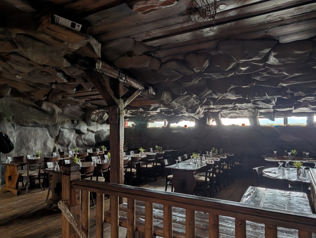 An interior view of a rustic dining area with wooden tables and chairs, surrounded by rock-like walls. The ceiling has a natural stone appearance, and there are small plants on the tables. Soft lighting illuminates the space, creating a cozy atmosphere.