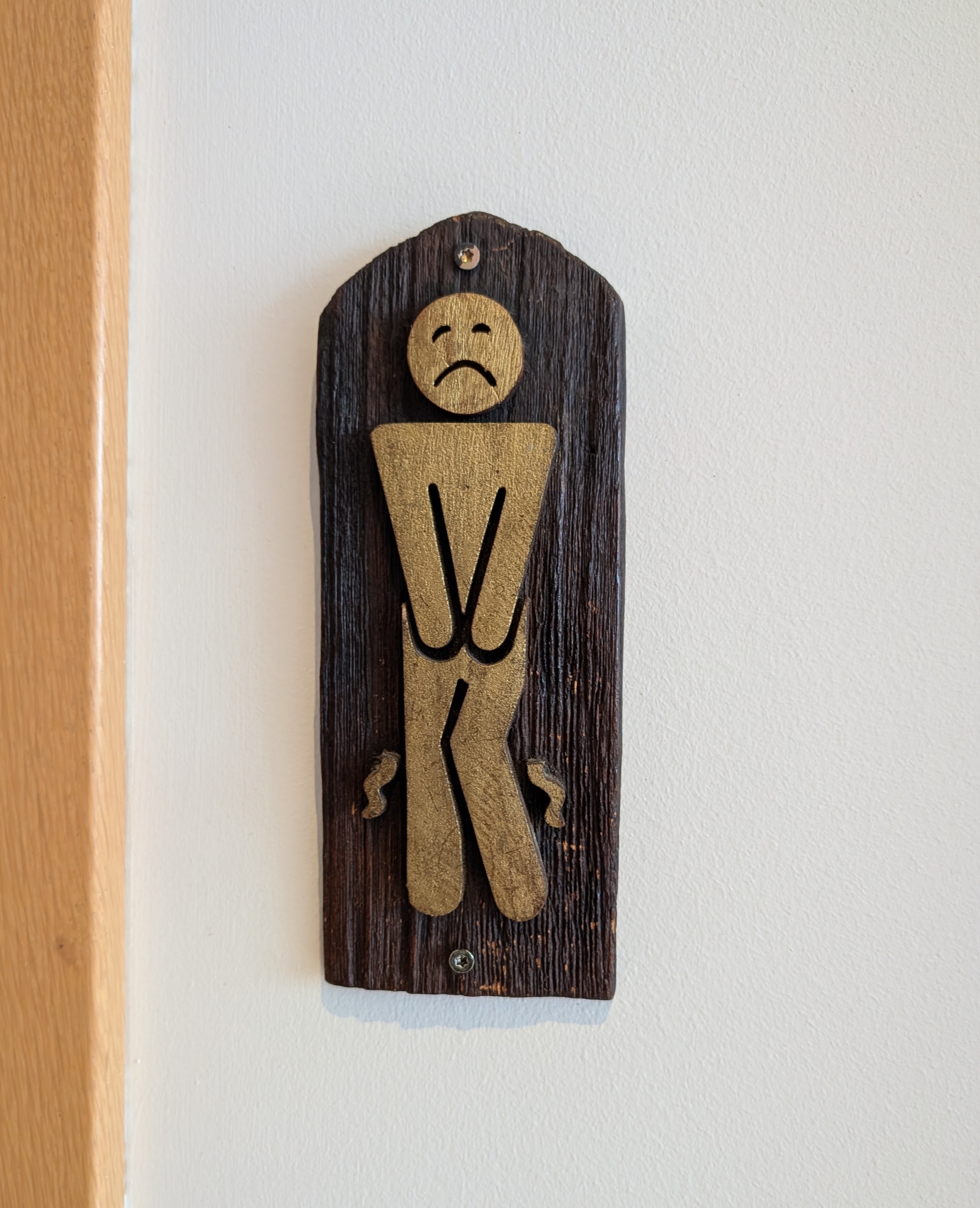 A wooden sign featuring a stylized figure with a sad expression, holding itself in a desperate posture, indicating a restroom.