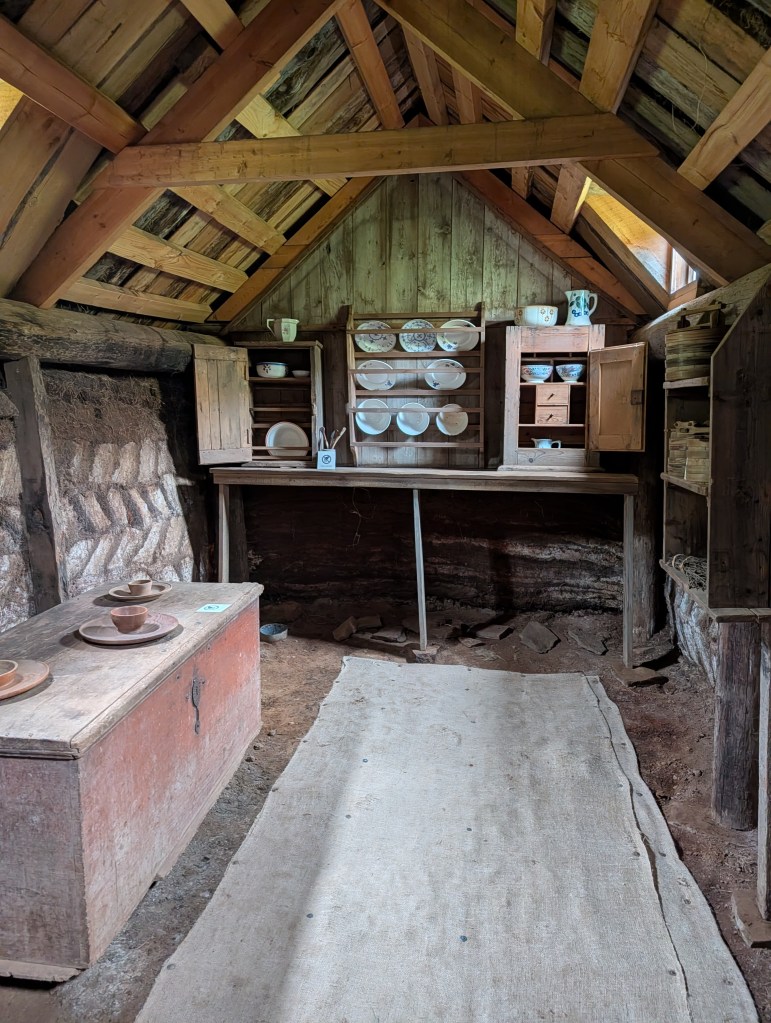 Interior view of a rustic kitchen with wooden beams, a table with pottery, and shelves displaying plates and cups.