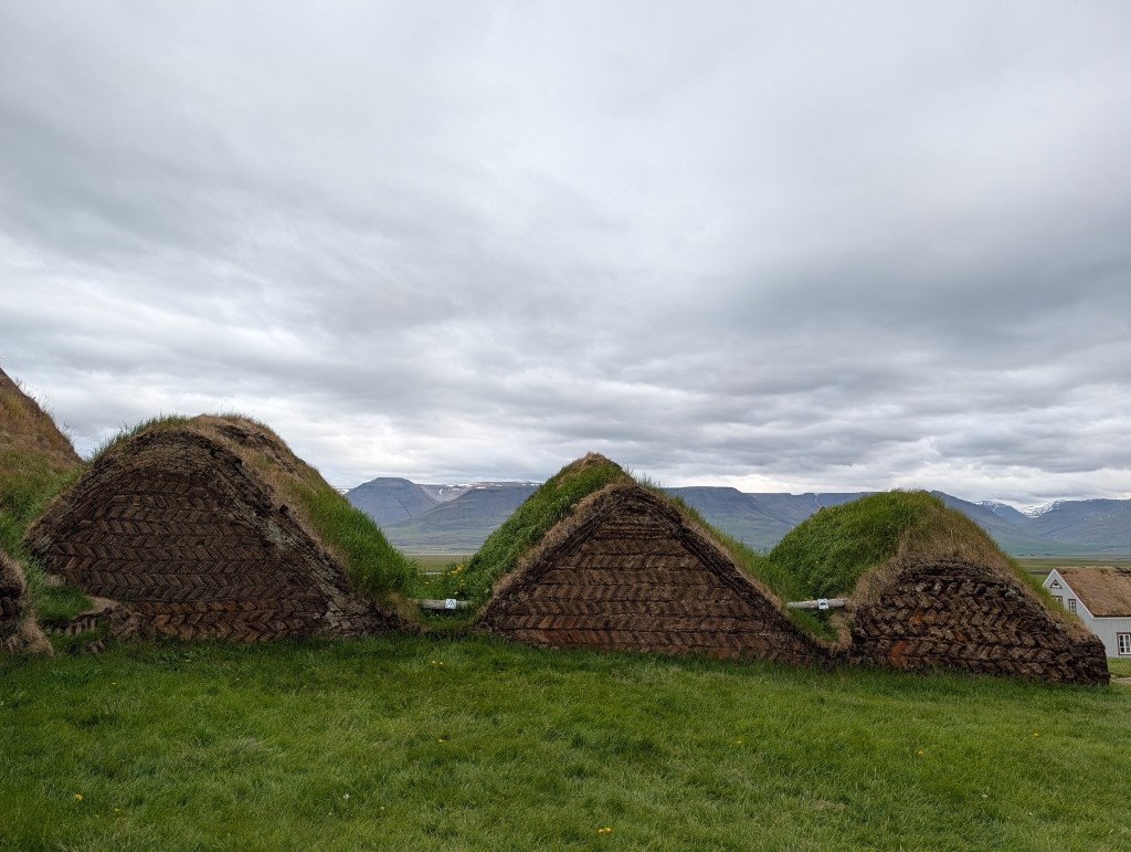 Traditional turf houses with grass roofs, set against a backdrop of rolling hills and cloudy skies in Iceland.