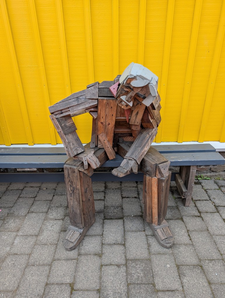 A wooden sculpture depicting a figure sitting on a bench, made from various pieces of reclaimed wood, set against a bright yellow wall.