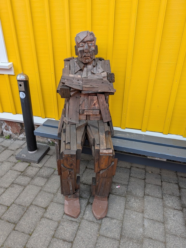 A wooden sculpture of a seated figure with arms crossed, made from various pieces of wood, set against a bright yellow wall.
