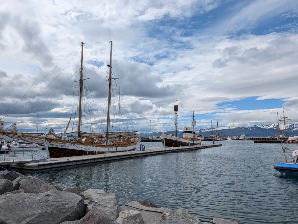 A scenic harbor with several boats docked by a wooden pier, against a backdrop of mountains and cloudy skies.