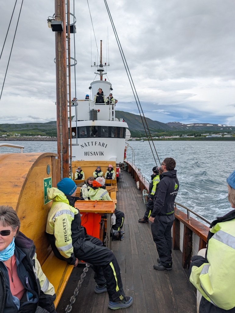Passengers and crew aboard the Nattfari boat in Husavik, surrounded by scenic coastal views and cloudy skies.