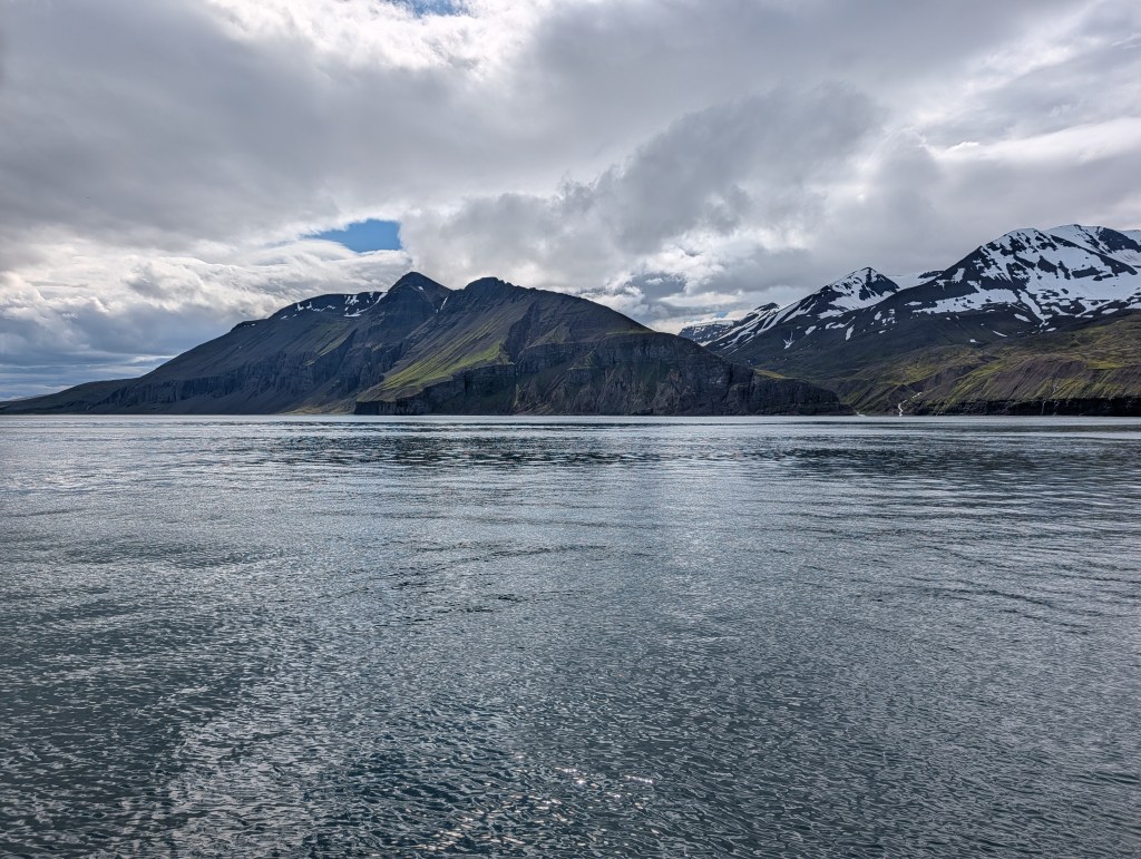 Scenic view of mountainous terrain with snow-capped peaks reflected in calm water under a cloudy sky.