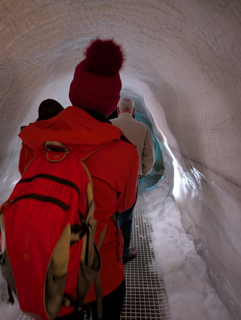People walking through a snow tunnel with icy walls, featuring a person in a red jacket and hat in the foreground.