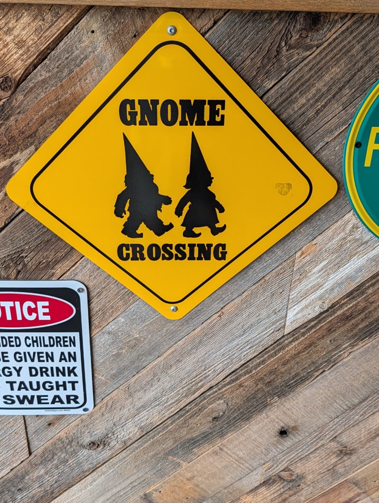A yellow traffic sign that reads 'Gnome Crossing' with silhouettes of two gnomes on it, mounted on a rustic wooden background.