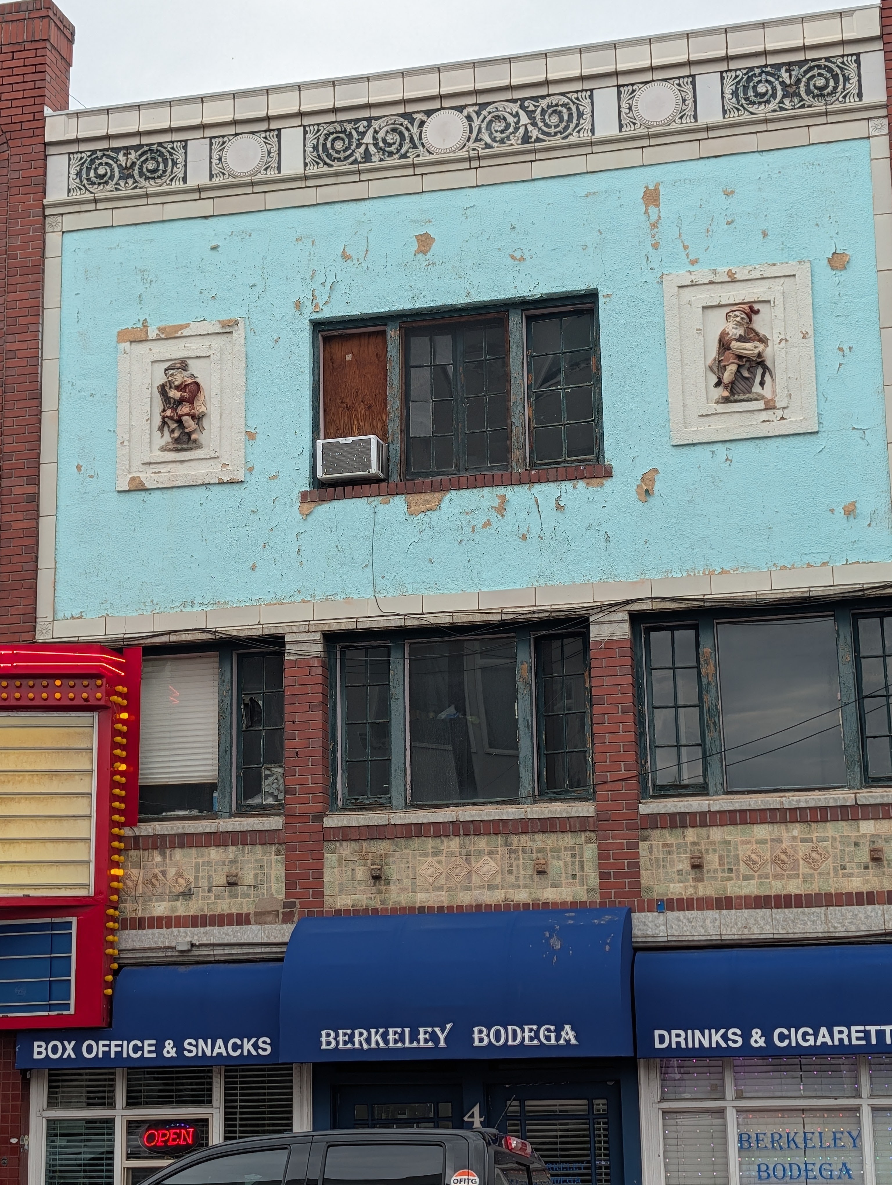 Front view of a building with peeling blue paint, featuring decorative reliefs on either side of the upper windows. The structure includes signs for 'Berkeley Bodega' and 'Box Office & Snacks.'