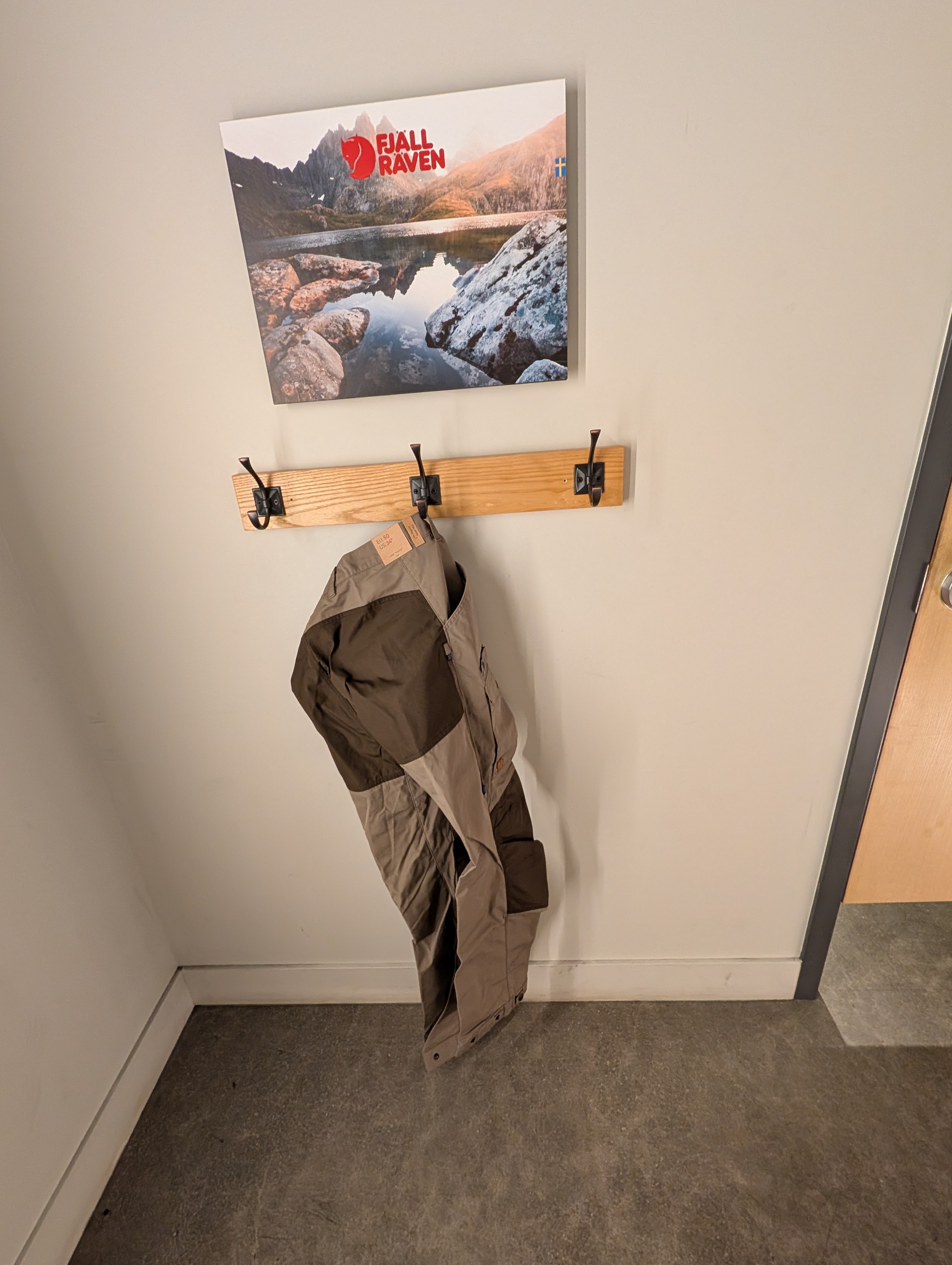 A Fjällräven poster featuring a scenic landscape hangs above a wooden coat rack with hooks, displaying a brown and gray jacket hanging from one of the hooks, in a minimalist indoor setting.