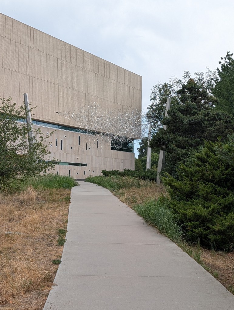 A paved walkway leading towards a modern building with a textured facade, surrounded by greenery and sparse trees.