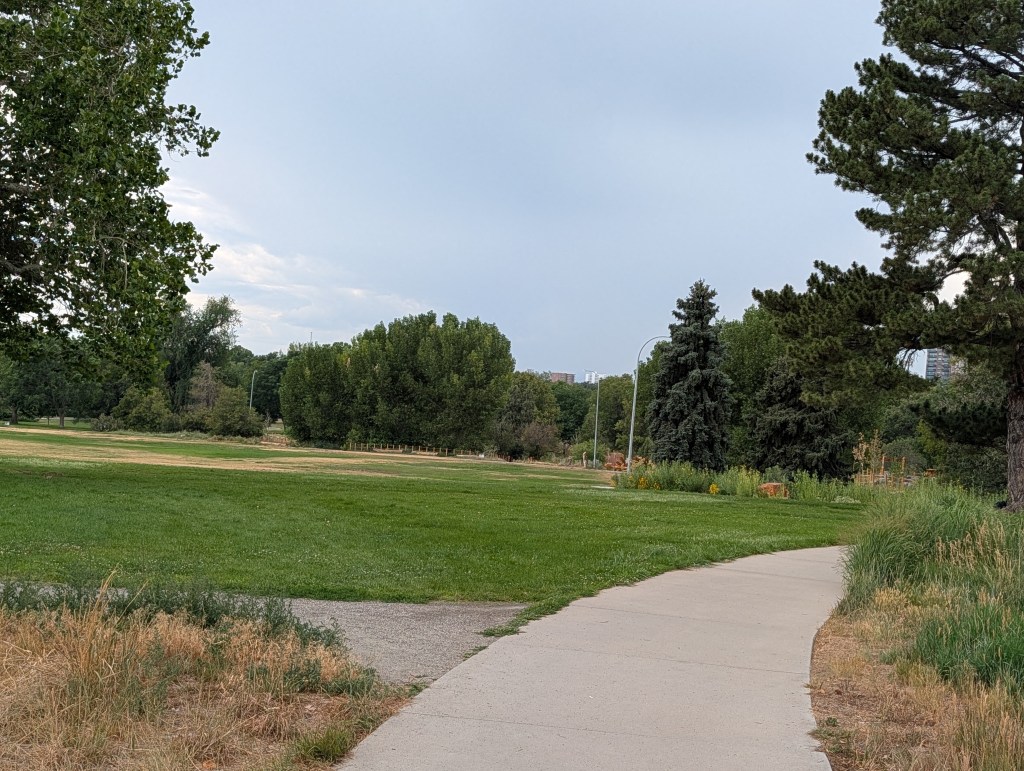 A peaceful park scene featuring a paved path winding through lush green grass and trees, with a cloudy sky in the background.