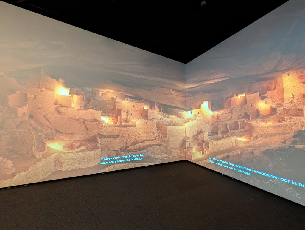A projection of ancient ruins at Mesa Verde, highlighting the effects of drought-fueled fires on the landscape, with bilingual captions in English and Spanish.