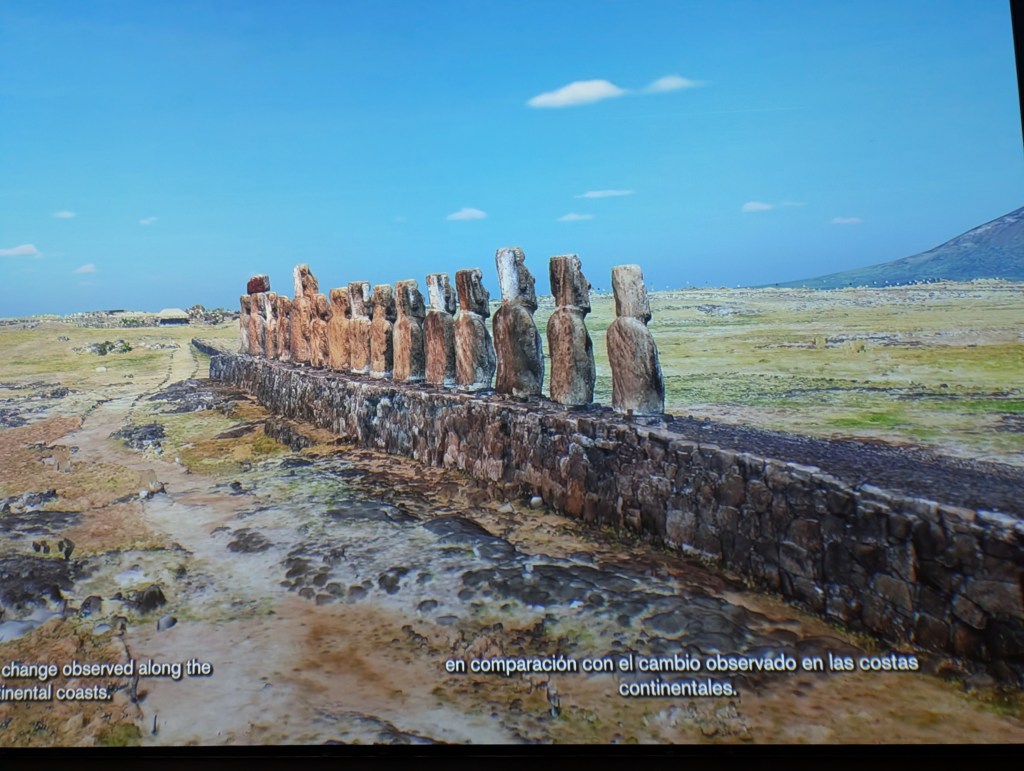 A row of ancient stone statues, known as Moai, standing on a stone platform against a clear blue sky, with an expansive landscape of grass and rocks in the background.