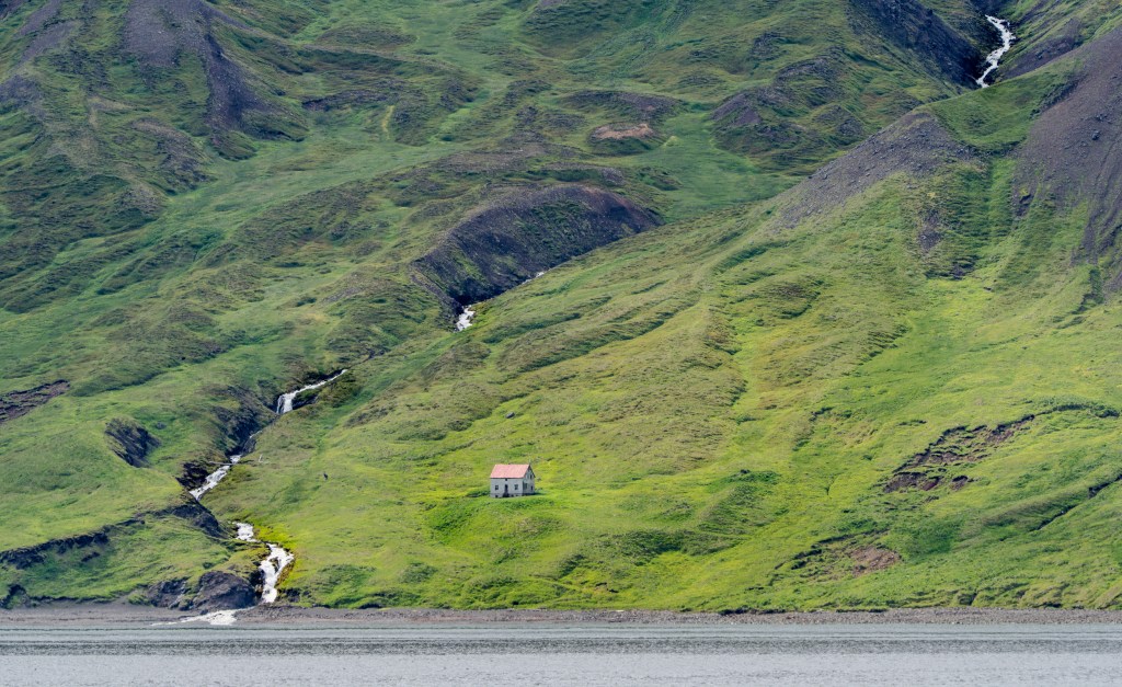 A small house with a red roof sits on a lush green hillside, surrounded by gentle slopes and small streams flowing down the landscape.